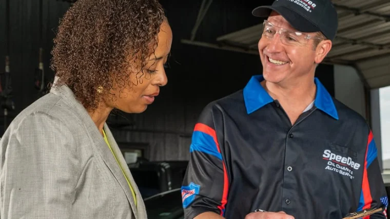 A mechanic in uniform holds a clipboard and speaks with a woman in a blazer inside an auto repair shop.