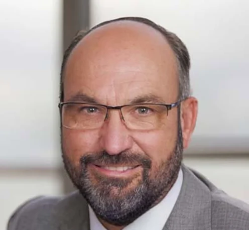 Ron Stilwell with glasses, a beard, and a bald head, wearing a gray suit and blue tie, smiling at the camera.
