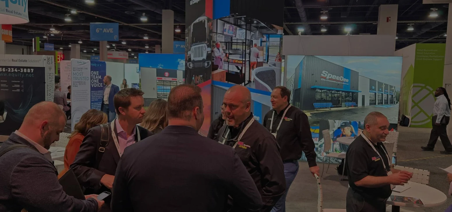 Group of people talking at an indoor trade show booth for SpeeDee Oil Change & Auto Service, with banners and informational materials visible.