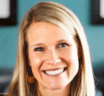 Stacey Pool with long blonde hair smiling, wearing gold earrings, in front of a blue background.