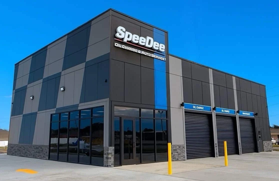 A modern SpeeDee Oil Change & Auto Service building with large windows and three service bays, set against a clear blue sky.